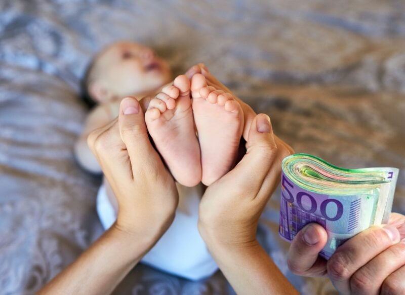 prikaz mamke koja drži bebu za nogice i pored je druga ruka sa novcem