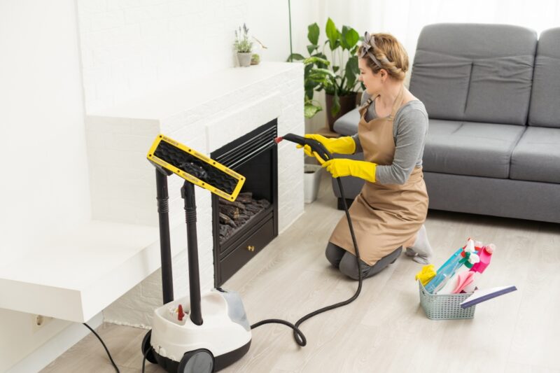 devojka koja se bavi profesionalnim čišćenjem drži usisivač u ruci i čisti kamin, pored nje kanta sa hemikalijama i proizvodima za čišćenje