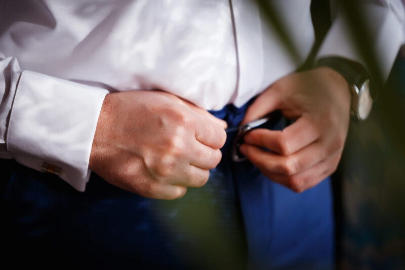 Muškarac u beloj košulji zakopčava crni kaiš na plavim pantalonama